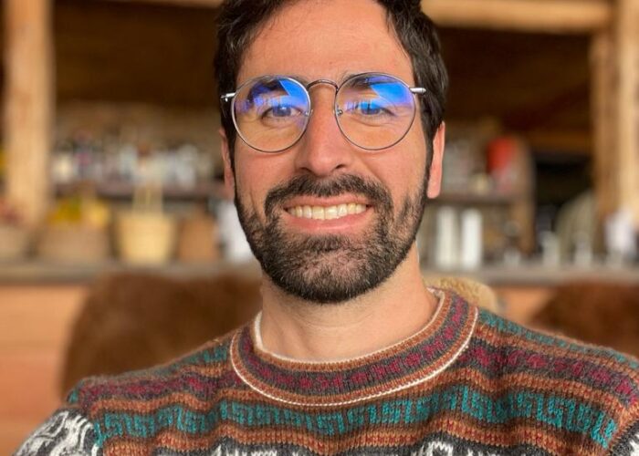 Homem com óculos redondos, barba e cabelos negros, vestido com blusa com desenho de lhamas dos Andes.